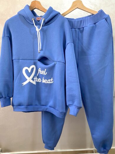La Survette d'allaitement, composée d'un sweat à capuche bleu avec un cœur blanc et le texte "feel the beat" en blanc sur le devant, ainsi que d'un pantalon de survêtement assorti, est suspendue à des cintres en bois.