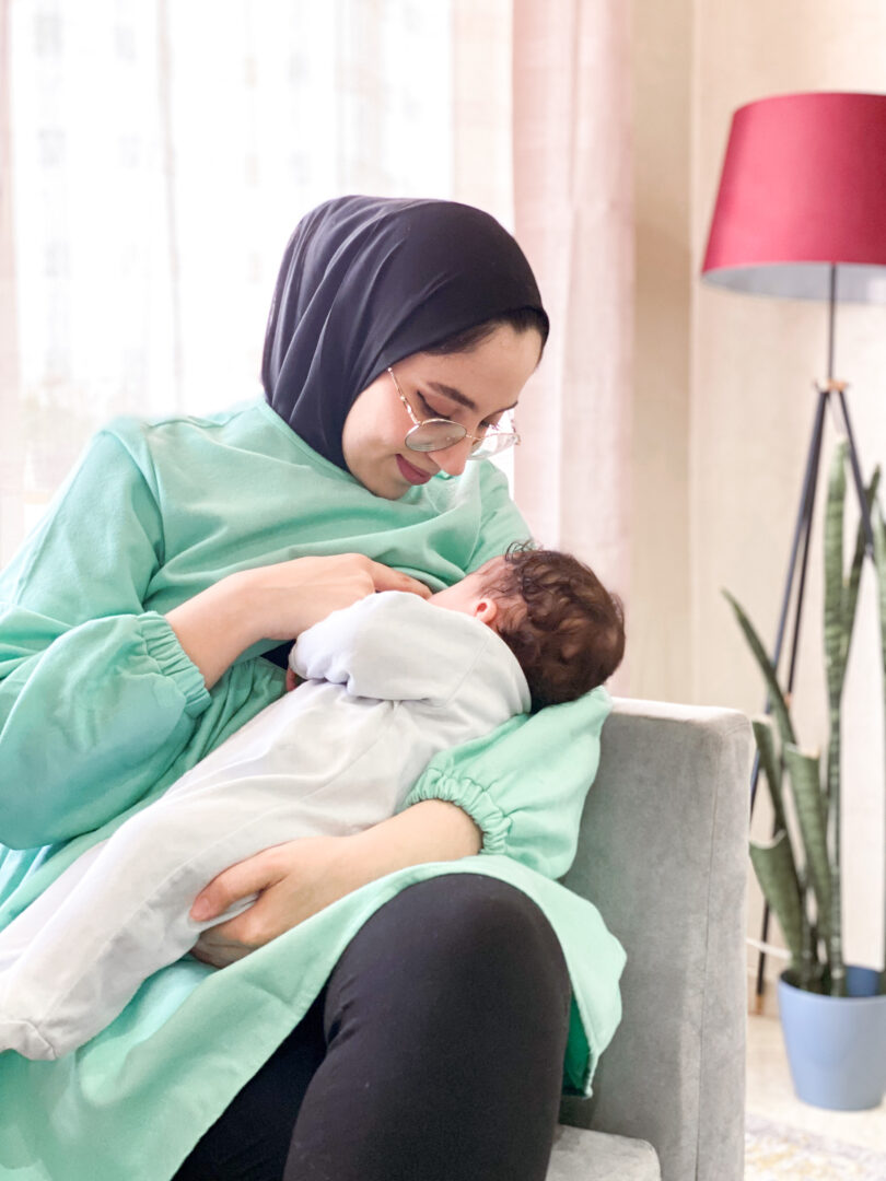 Une femme portant des lunettes et un foulard noir allaite un bébé assise sur une chaise gris clair. Elle porte la Robe 2 en 1 : De grossesse et d’allaitement en vert clair, associée à un pantalon noir. Une grande plante et un abat-jour rouge se dressent en arrière-plan à côté d’un rideau.