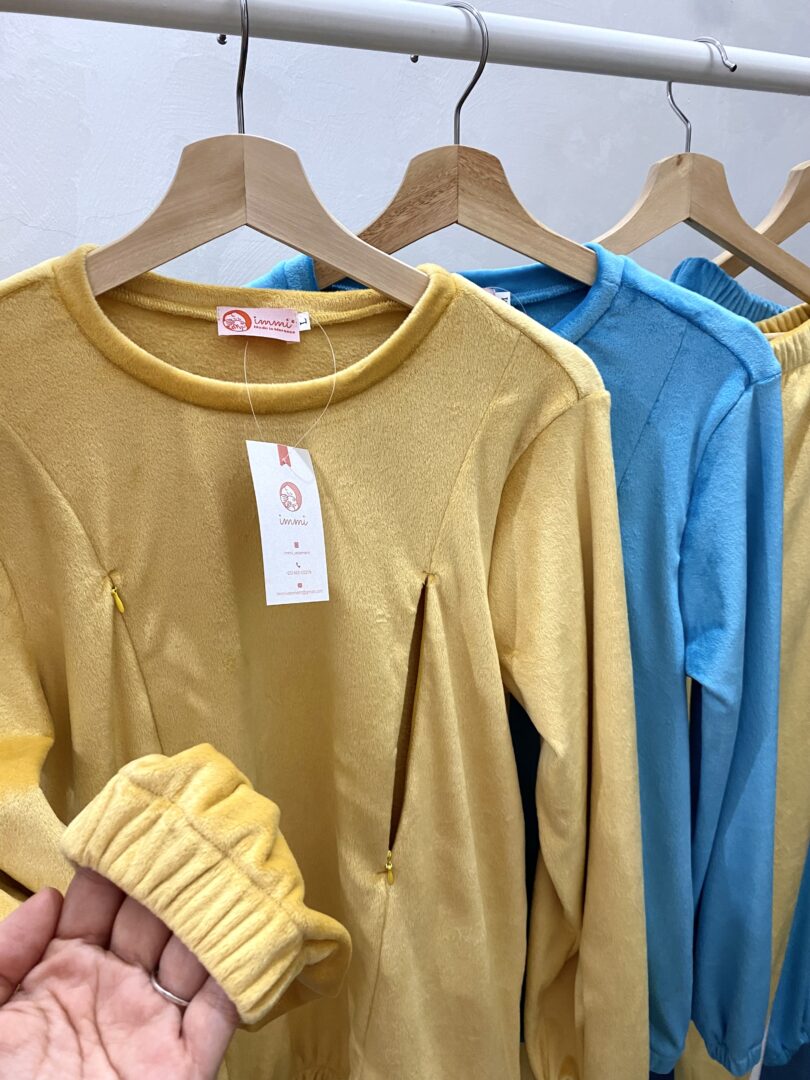 Une main tenant une manche jaune de Pyjamas d’allaitement, exposant un portant de vêtements douillets. Le présentoir comprend une série de hauts et de pantalons à manches longues en jaune et bleu. Les vêtements portent des étiquettes attachées à des cintres en bois. L’arrière-plan est un mur blanc uni.