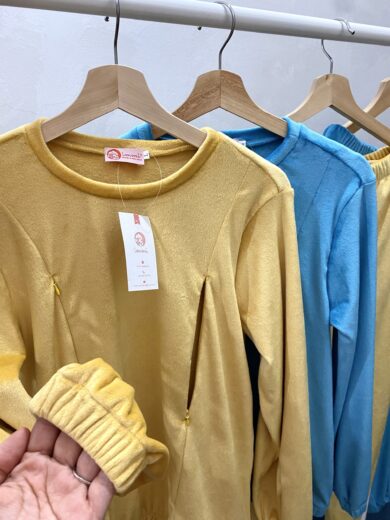Une main tenant une manche jaune de Pyjamas d’allaitement, exposant un portant de vêtements douillets. Le présentoir comprend une série de hauts et de pantalons à manches longues en jaune et bleu. Les vêtements portent des étiquettes attachées à des cintres en bois. L’arrière-plan est un mur blanc uni.
