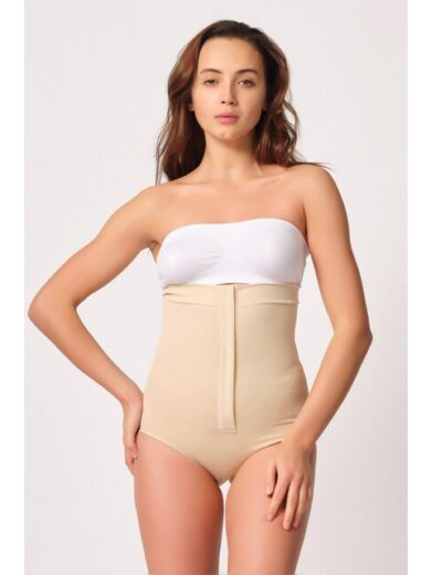 Une femme portant le Corset Culotte Attaché Devant 2088 en beige et un haut bandeau sans bretelles blanc se dresse sur un fond uni. Elle a une main sur sa hanche et l'autre détendue à ses côtés. Elle a les cheveux longs et ondulés et une expression neutre.