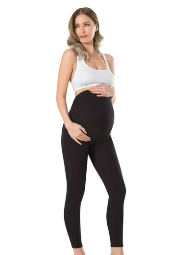 Une personne enceinte se tient sur un fond blanc uni, portant un soutien-gorge de sport blanc et un Legging de grossesse en coton 665 noir. Elle berce doucement son baby bump avec les deux mains, regardant vers la gauche avec un léger sourire.