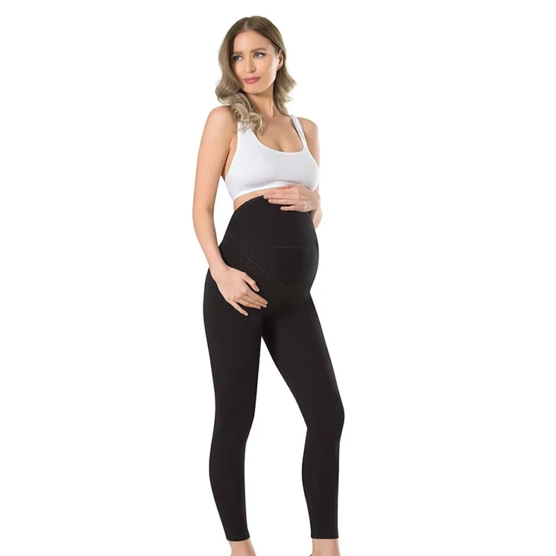 Une personne enceinte se tient sur un fond blanc uni, portant un soutien-gorge de sport blanc et un Legging de grossesse en coton 665 noir. Elle berce doucement son baby bump avec les deux mains, regardant vers la gauche avec un léger sourire.