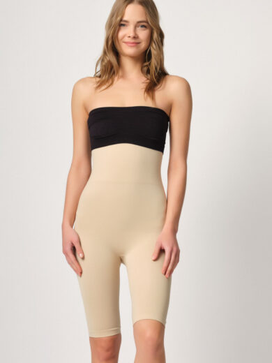Une femme aux longs cheveux blonds ondulés se tient devant un fond uni, portant le Corset gaine court moulant taille haute 2014. Ce shapewear bustier taille haute de couleur nude s'étend jusqu'à mi-cuisse, avec la partie supérieure en noir. Elle sourit et semble détendue.